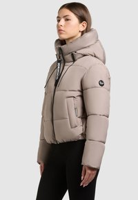 Lekka beżowa kurtka puchowa z wysokim kołnierzem, przednim zamkiem błyskawicznym i czarnymi akcentami logo. Posiada boczne kieszenie oraz miękką, pikowaną fakturę.