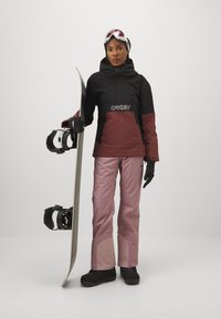 Snowboardeur en veste noire et bordeaux, pantalon rose, gants noirs, tenant un snowboard. Présente des lunettes de protection teintées de rose et des boots noires.