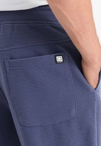 Tmavě modré tepláky s žebrovanou texturou, s kapsou na zadní straně a čtvercovou nášivkou v horním rohu.