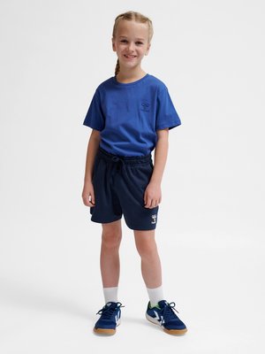 Lächelndes Mädchen mit geflochtenem Haar trägt ein blaues T-Shirt, marineblaue Shorts, weiße Socken und blaue Turnschuhe, steht vor einem schlichten weißen Hintergrund.