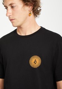 Sort bomulds T-shirt med et cirkulært orange grafisk design med stråler og et dråbe symbol på brystet. Korte ærmer og rund hals.