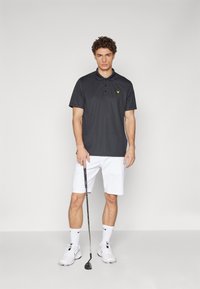 Dark grey polo shirt with a yellow logo, short sleeves, and a collar. Paired with white shorts and golf shoes, holding a golf club.