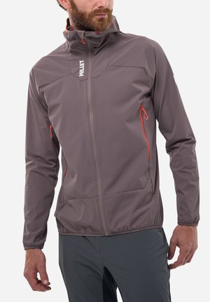 Homme portant une veste violette à fermeture éclair avec des tirettes orange et un col montant, debout les mains le long du corps devant un fond uni.