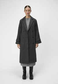 Un long manteau en laine gris au design double boutonnage, avec de larges revers et des manches surdimensionnées, associé à une robe en maille grise et des bottines noires.