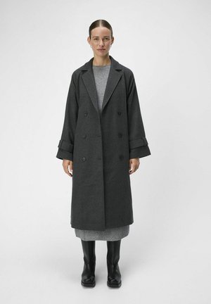 Un long manteau en laine gris au design double boutonnage, avec de larges revers et des manches surdimensionnées, associé à une robe en maille grise et des bottines noires.