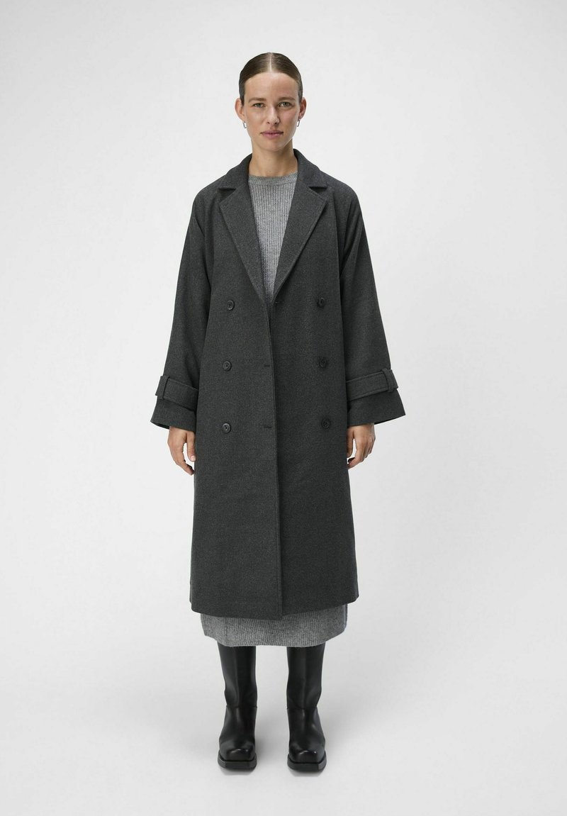 Un long manteau en laine gris au design double boutonnage, avec de larges revers et des manches surdimensionnées, associé à une robe en maille grise et des bottines noires.