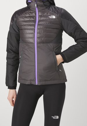Woman wearing a black and dark gray North Face jacket with a purple zipper and black North Face leggings against a plain background.