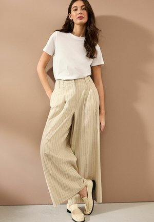 Beżowe spodnie szerokie w paski z przodu, zestawione z gładką białą koszulą z krótkim rękawem i jasnymi butami wsuwanymi.