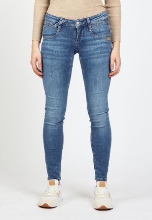 Frau trägt hellblaue Skinny-Jeans und weiße Sneakers, steht vor einem schlichten weißen Hintergrund.