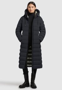 Cappotto lungo imbottito di un nero intenso, dotato di cappuccio, chiusura con zip e tasche laterali; esterno trapuntato e fodera interna a contrasto.