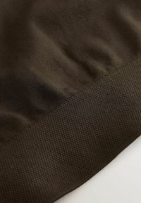 Tumepruun kangas, millel on sujuv ülemine osa ja tekstuuriga kudumise muster allosas. Õmbluse detail on nähtav ääres.