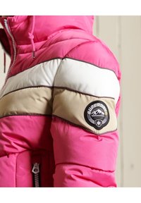 Lyserød pufferjakke med hvide og beige vandrette striber. Inkluderer et rundt sort logo-patch og en lynlåslomme med en sølvlynlås.