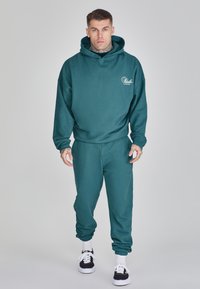 Mörk teal hoodie och jogger-set i mjukt tyg, med en avslappnad passform, ärmmuddar och muddar vid anklarna, samt en logodetalj på framsidan.