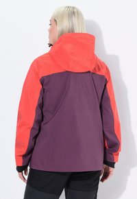 Giacca impermeabile viola e rossa con cappuccio, caratterizzata da un design colorato a blocchi e bordi neri sui polsini. Texture liscia, vestibilità rilassata.