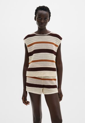 Nő ujjatlan horgolt felsőt és hozzáillő rövidnadrágot visel, barna, narancssárga és krémszínű vízszintes csíkokkal, fehér háttér előtt áll.