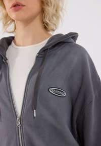 Siva zip-up dukserica s kapuljačom i crnim ovalnim logom. Ima mekanu teksturu i složena je preko bijele rebraste majice.