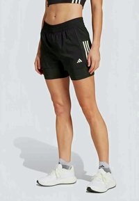 Ženska v črnih športnih kratkih hlačah Adidas, belih nogavicah Adidas in belih tekaških čevljih, ki stoji ob enobarvni podlagi.