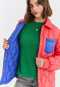 Žena v korálově prošívané bundě s modrým kapsou a podšívkou, zeleném svetru, šedých džínách a zlatém přívěsku na krku.