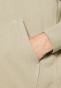 Felpa leggera beige con tasca frontale, tessuto strutturato e polsini a coste. Una mano appoggiata nella tasca evidenzia la vestibilità del capo.