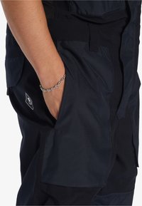 Zwarte technische broek met zakken, met een zachte textuur en een slim fit ontwerp. Kettingarmband zichtbaar om de pols. Logo detail aanwezig.