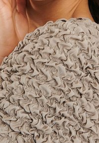 Beige textiel met een geplooide, gerimpelde uitstraling. Het oppervlak bevat georganiseerde plooien, wat een dimensionale en gelaagde uitstraling creëert.