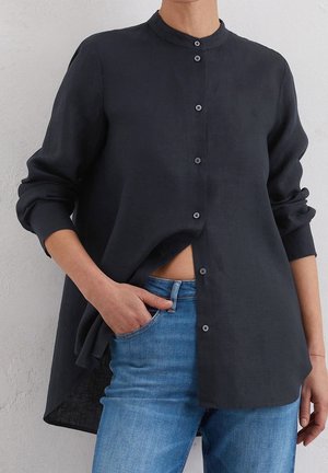 Persoon draagt een donker overhemd met opgerolde mouwen en een blauwe spijkerbroek, hand in de zak, staand tegen een lichte muur.