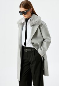 Abrigo de lana gris con un collar de piel sintética, llevado sobre una camisa blanca y una corbata negra, combinado con pantalones negros y accesorios que incluyen gafas de sol.