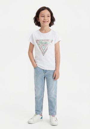 Kind mit schulterlangem dunklem Haar, trägt ein weißes Guess-T-Shirt, hellblaue Jeans und weiße Sneakers, lächelt und steht mit einer Hand in der Tasche.