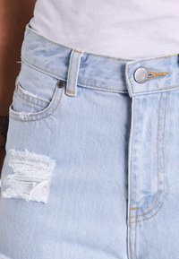 Jean en denim bleu clair à taille haute, avec des détails usés sur la cuisse gauche et une fermeture à bouton en métal, comportant un design classique à cinq poches.