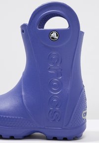 Bota de goma morada con superficie texturizada, diseño redondeado, etiqueta de logo y marca "crocs" en relieve a un lado. Incluye un asa para facilitar su uso.
