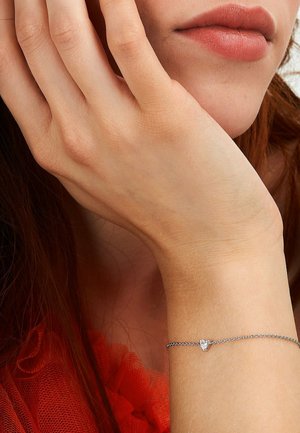 Close-up of a woman's hand resting on her cheek, wearing a delicate silver bracelet with a small heart charm.