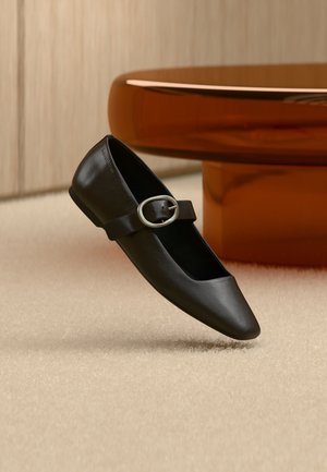 Chaussure plate Mary Jane en cuir noir avec boucle argentée, exposée sur un tapis beige près d'un pied de table rond marron.