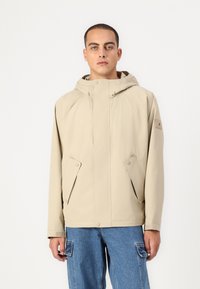 Beige jakke med hette, laget av glatt materiale. Har trykknapper, to frontlommer og et logo-patch på ermet.