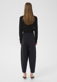 Donna con lunghi capelli castani lisci che indossa un top nero a maniche lunghe, pantaloni blu navy a vita alta e sandali neri con tacco, girata di spalle.