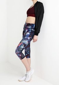 Højtaljede capri-bukser i et livligt blomsterprint, lavet af strækbart stof. Kombineret med en maroon crop top og en sort letvægtsjakke. Hvide sneakers.