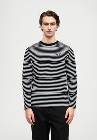 Fiatal férfi áll, fekete-fehér vízszintes csíkos hosszú ujjú pólót visel, amelyen szöveg található a mellkasán, és fekete nadrág van rajta, sima fehér háttér előtt.