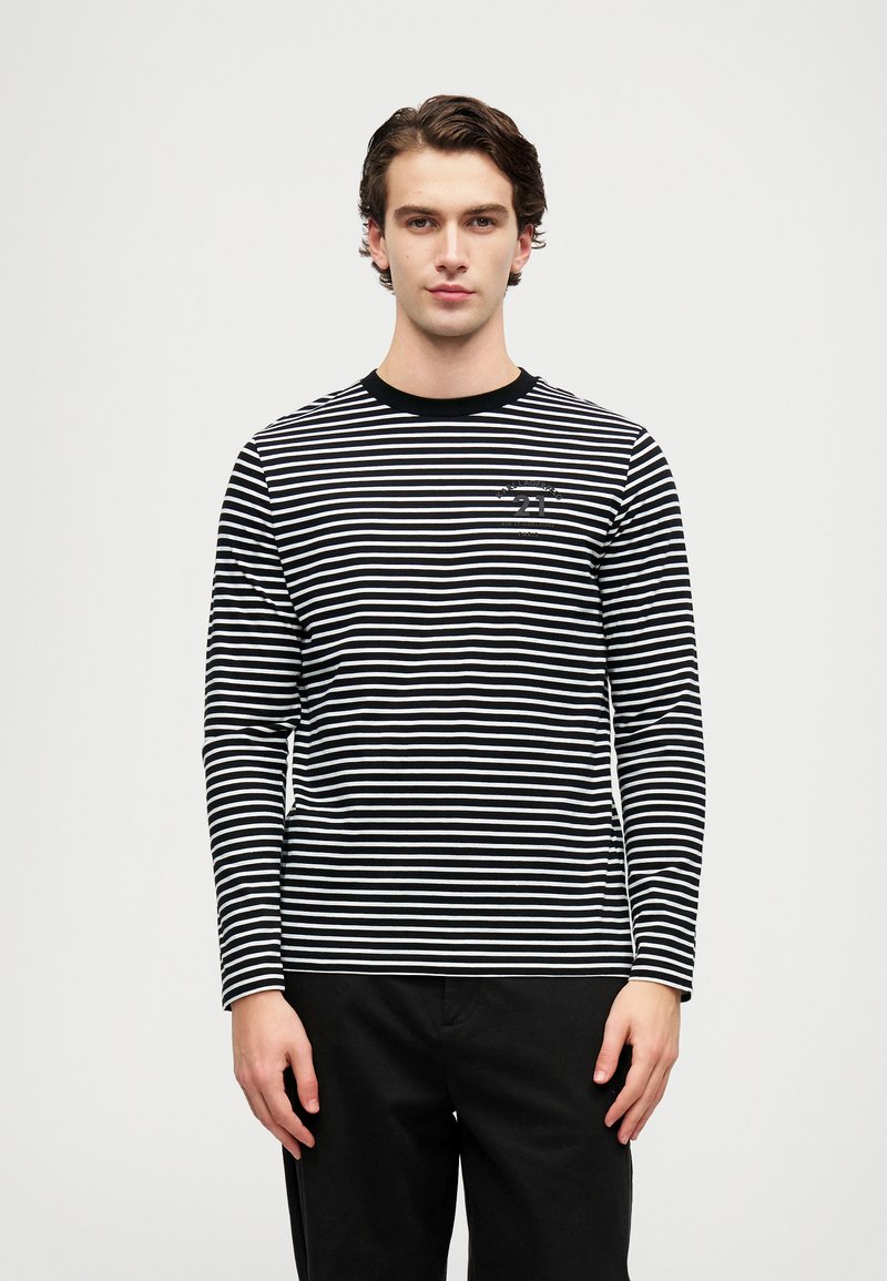 Fiatal férfi áll, fekete-fehér vízszintes csíkos hosszú ujjú pólót visel, amelyen szöveg található a mellkasán, és fekete nadrág van rajta, sima fehér háttér előtt.