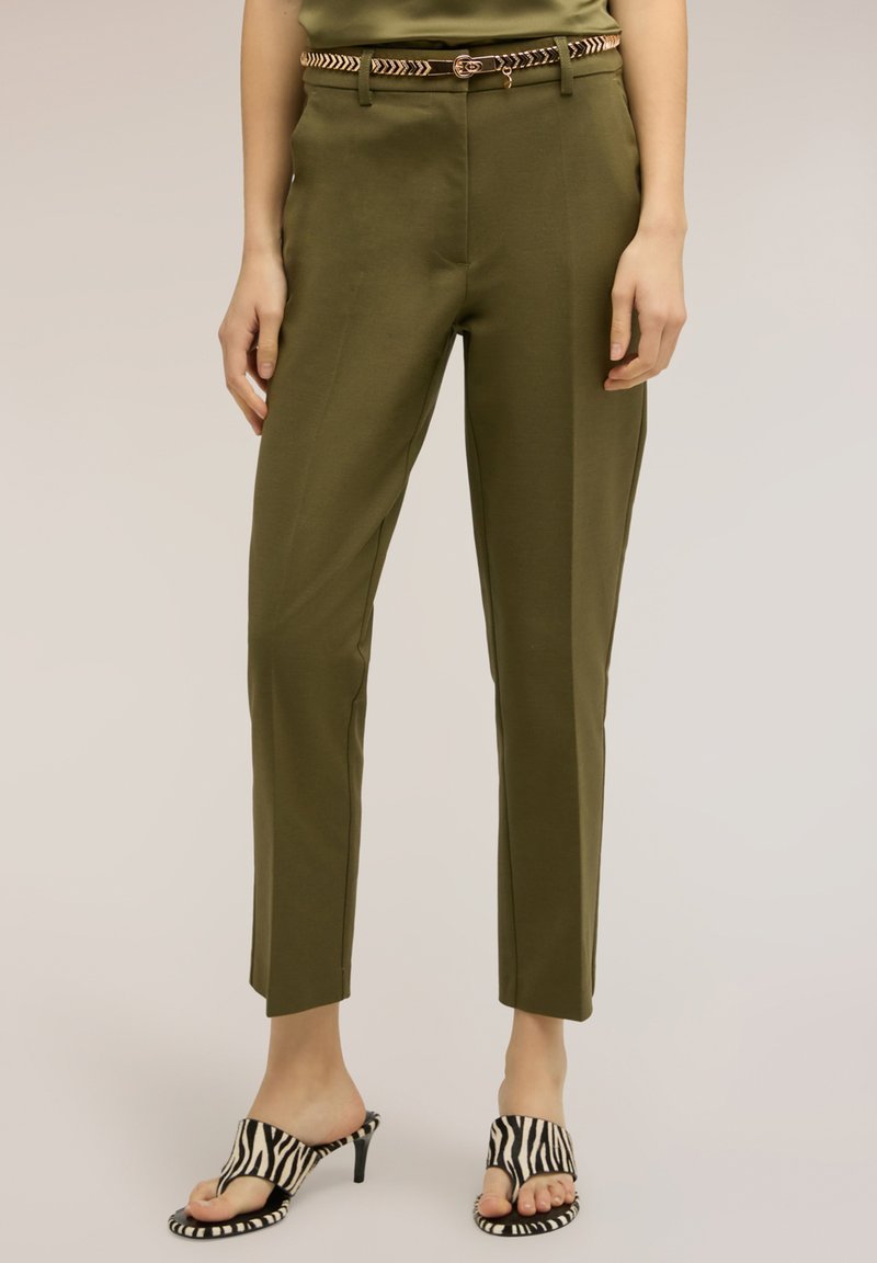 Mujer con pantalones entallados verde oliva y un cinturón delgado con estampado, y sandalias de tacón con rayas de cebra, de pie frente a un fondo liso.