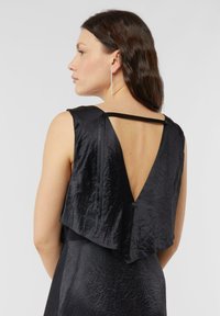 Czarna sukienka z fakturowanej tkaniny, z dekoltem w kształcie litery V na plecach i bez rękawów. Podkreślona wąskim paskiem i przepływającym wielowarstwowym detalem na plecach.