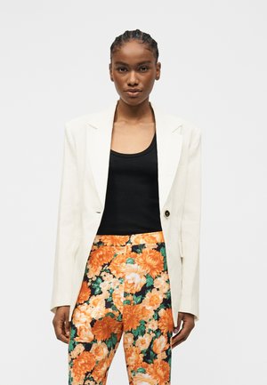 Vrouw met gevlochten haar die een witte blazer, zwarte top en hooggetailleerde broek met oranje bloemmotief draagt, staand tegen een effen achtergrond.