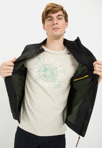 Schwarze Außenjacke mit Druckknopfverschluss, grüne Futter. Beiges T-Shirt darunter mit einem kreisförmigen grafischen Design in Aqua und Weiß.