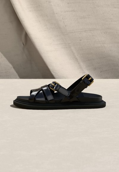 Μαύρο δερμάτινο σανδάλι με δύο χρυσά αγκράφες, λουριά που διασταυρώνονται και επίπεδη σόλα, παρουσιάζεται πάνω σε ουδέτερο υφασμάτινο φόντο.