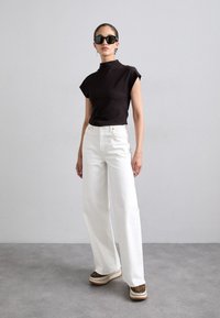 Haut ajusté noir, jeans blancs à jambes larges et chaussures à plateforme avec un motif léopard. Les accessoires incluent des lunettes de soleil et des boucles d'oreilles. Arrière-plan minimaliste.
