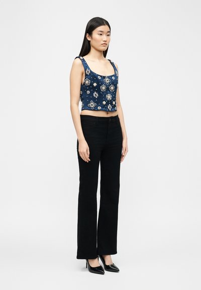 Γυναίκα που φοράει μπλε αμάνικο crop top με λουλουδένιες διακοσμήσεις, μαύρο ψηλόμεσο παντελόνι και μαύρα ψηλοτάκουνα παπούτσια, όρθια πλάγια.