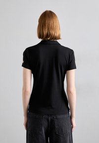 Pólo preto com mangas curtas, colarinho e um subtil detalhe dourado no ombro esquerdo. Textura suave e corte ajustado.