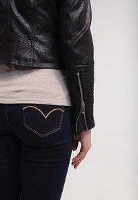 Zbliżenie na osobę w czarnej skórzanej kurtce z zapięciami na rękawach, beżowej koszuli i ciemnoniebieskich dżinsach z żółtym przeszyciem na tylnej kieszeni.