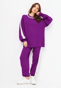 Sweatshirt en tricot violet avec des accents de rayures blanches, associé à un pantalon ample assorti. Les deux pièces présentent des poignets et un ourlet côtelés. Des baskets blanches complètent le look.