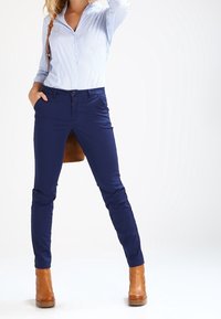 Marineblauwe slim-fit broek met een gladde textuur, gecombineerd met een lichtblauw gestreept shirt. Bruine leren laarzen met hak en een bruine handtas.