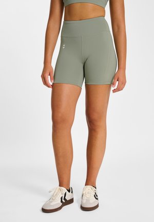 Vrouw draagt lichtgroene, hooggetailleerde sportshorts en een bijpassende crop top, gecombineerd met witte en zwarte sneakers, staand op een witte vloer.