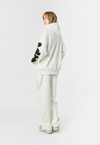 Sudadera blanca de gran tamaño con un diseño negro en la manga, combinada con pantalones blancos y zapatos de texturizados gris claro, con un detalle de cadena.